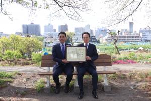 寄贈（リニューアル）されたまちなかベンチに座る、橋本太樹代表取締役社長と富岡市長です。感謝状を手に笑顔で写っています。