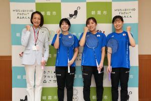 交付式の記念写真です。市長と出場予定選手がバドミントンのラケットを手にガッツポーズをとっています。