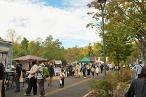 アサカストリートテラスの様子です。シンボルロードの色づく木々の下で、さまざまな出店が立ち並び、参加者が買い物をしています。