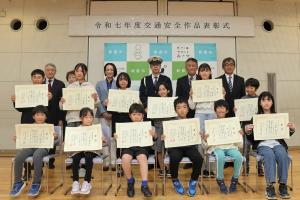 令和7年度交通安全作品表彰式の記念写真です。表彰された児童が表彰状を手にし並んでおり、その後ろに市長等の来賓が並んでいます。