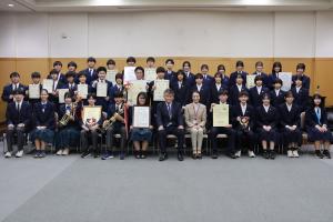 報告会の記念写真です。松下市長と二見教育長を中心にして、大会へ出場した中学生が賞状やトロフィー等を手に写っています。