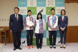 表敬訪問の写真です。二見教育長と松下市長が両脇に立ち、泉コーチ、佐藤選手、原田選手がフライングディスクとメダルを手に写っています。