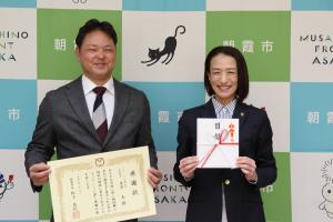 感謝状贈呈式の記念写真です。朝霞市のバックパネルの前に代表取締役と市長がそれぞれ、感謝状と目録を手にうつっています。