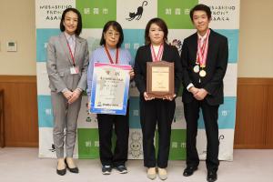 表敬訪問の記念写真です。朝霞市のバックパネルの前で監督・選手が賞状とメダルを手にして、市長と一緒に写っています。