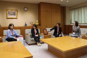 表敬訪問の様子の写真です。市長・監督・選手が椅子に座り談笑しており、監督がメダルと賞状の内容を説明しています。