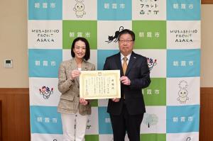 感謝状贈呈式の記念写真です。朝霞市のバックパネルの前で、市長と代表取締役社長が感謝状を手にして写っています。
