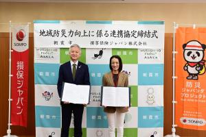 協定締結式の記念写真です。朝霞市のバックパネルと損保ジャパンののぼりばたの前で協定書を手にした損保ジャパン埼玉支店長と市長が写っています。
