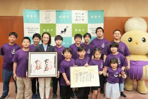 表敬訪問の記念写真です。松下市長とこども力士の皆さんが感謝状と横綱の手形色紙を手に写っています。回しをしめたぽぽたんもいます。