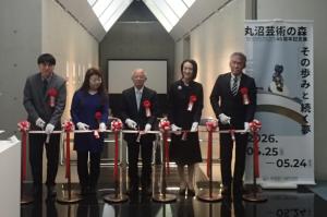 丸沼芸術の森コレクション展のオープニングセレモニーの際のテープカットの写真です。会場入り口に設置された紅白のテープを松下市長をはじめとした関係者がハサミを入れ、切ろうとしています。