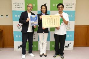 表敬訪問の記念写真です。表彰状を手にして松下市長と朝霞駅前商店会の代表者2人が笑顔で写っています。